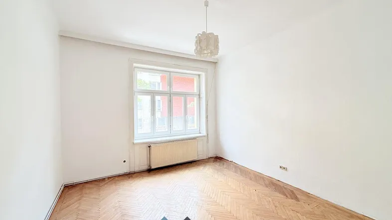 Helles Zimmer mit klassischem Fischgrätparkett, hohen Decken und einem großen Fenster.