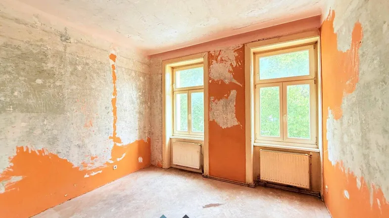 Unrenovierter Raum mit abgeblätterter Farbe an den Wänden und zwei Fenstern, bereit zur Sanierung.