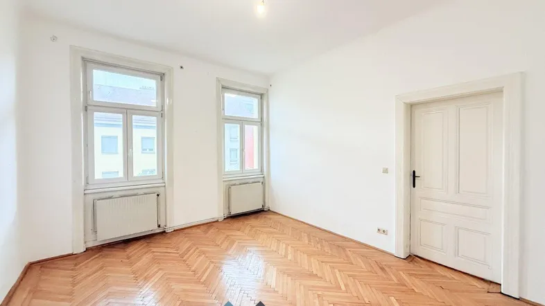 Helles, leeres Zimmer mit Fischgrätparkett und hohen Fenstern, bereit zur individuellen Gestaltung.