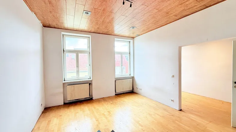 Helles Zimmer mit Holzdeckenverkleidung, Parkettboden und zwei Fenstern, bereit zur Renovierung.