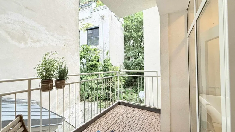 Heller Balkon mit Holzbodenfliesen, Metallgeländer und Blick ins Grüne der Umgebung.