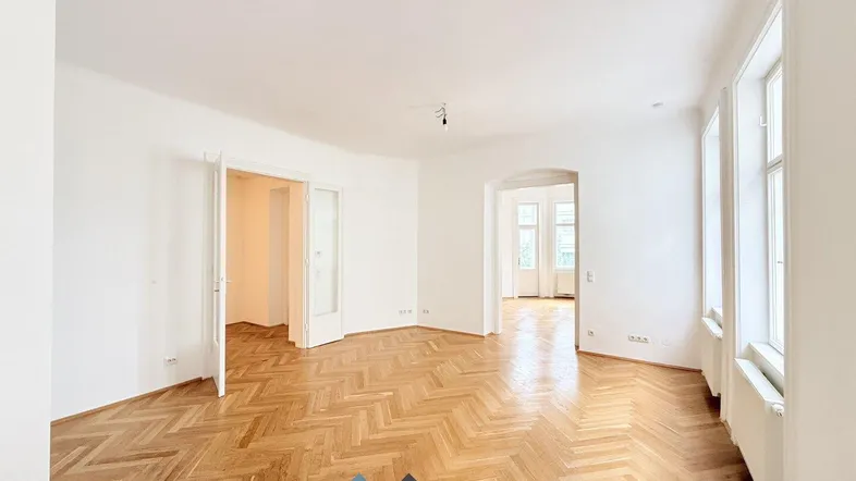 Helles Zimmer mit Fischgrätparkett und hohen Fenstern, das viel Tageslicht hereinlässt.