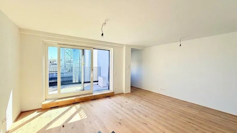 Heller, leerer Wohnraum mit Holzboden und Zugang zu einem Balkon.