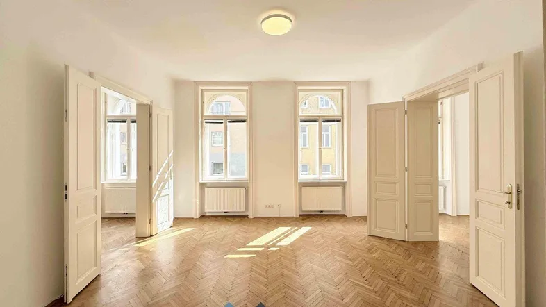 Helles, leeres Zimmer mit Fischgrätparkett und hohen Flügeltüren, das viel Tageslicht hereinlässt.