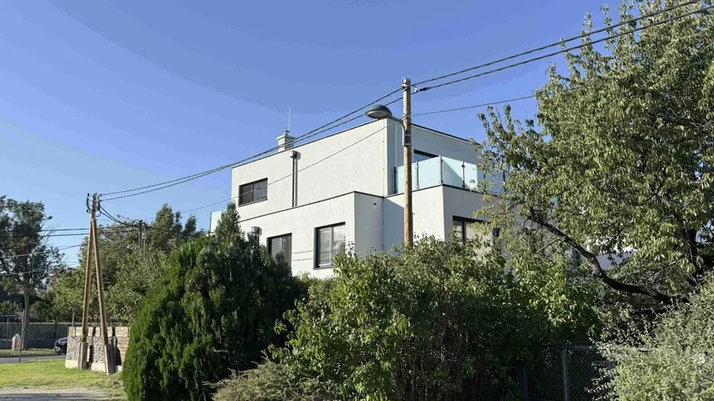 Modernes weißes Gebäude mit Flachdach und Balkon, umgeben von Bäumen und Büschen unter blauem Himmel.