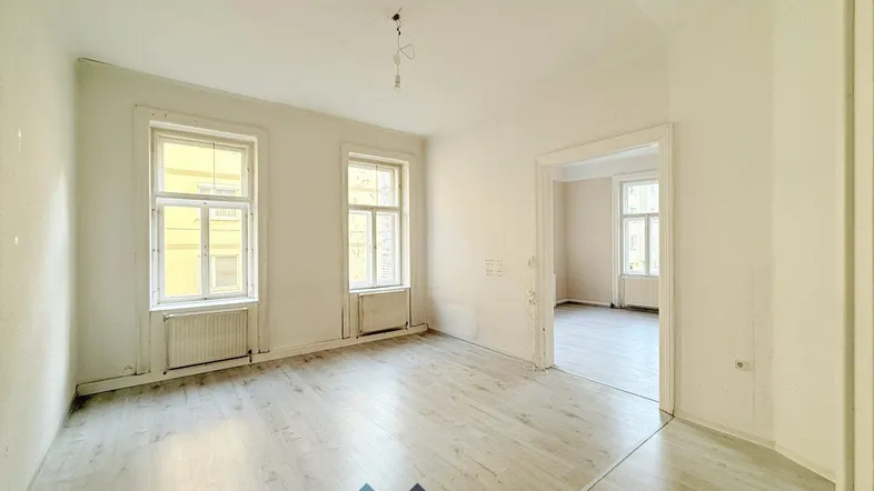 Helles, leeres Zimmer mit Laminatboden und zwei Fenstern, das auf einen weiteren Raum blickt.