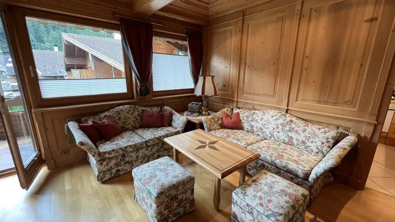 Gemütliches Wohnzimmer im alpinen Stil mit Holzvertäfelung, floralen Sofas und Blick ins Grüne.