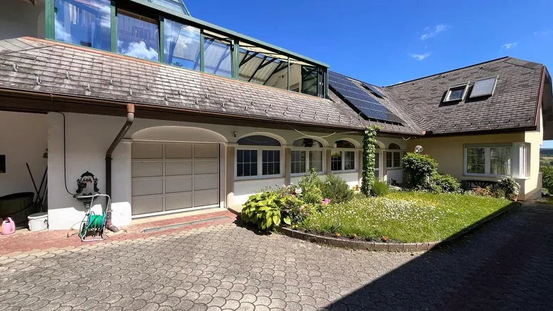 Ansicht des Hauses mit gepflasterter Auffahrt, Garage und Wintergarten unter blauem Himmel.