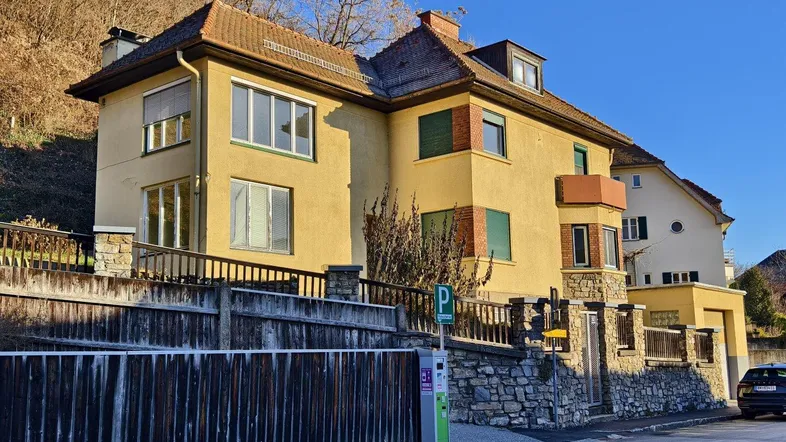 Gelbe Fassade eines Altbaus mit Ziegeldach und Steinsockel an einer Straße.