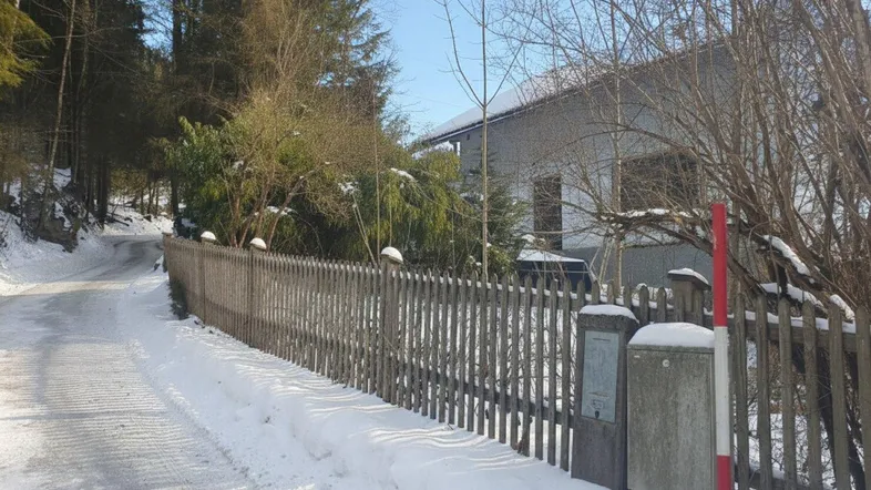 Winterliche Szene mit einem Holzzaun entlang einer verschneiten Straße und dem Haus im Hintergrund.