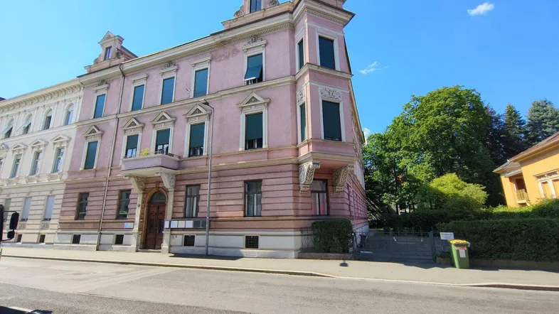 Historisches Gebäude mit rosa Fassade, grünen Fensterläden und dekorativen Elementen an einer Straßenecke.