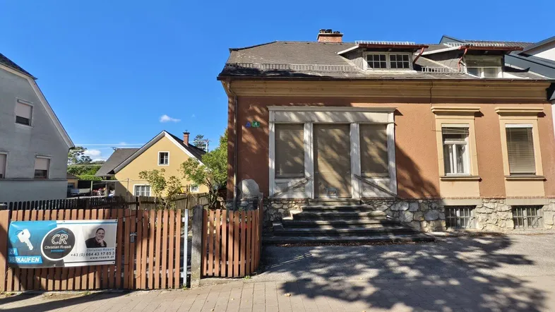 Historisches Gebäude mit Putzfassade und einem "Zu verkaufen"-Schild im Vorgarten.