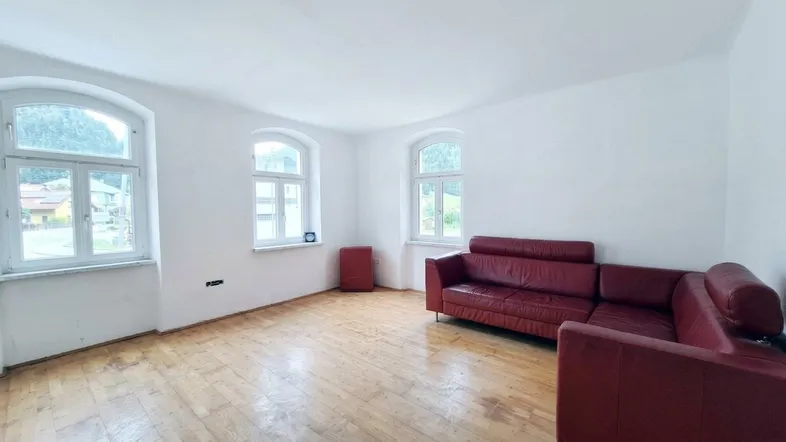 Helles Wohnzimmer mit Holzboden und zwei Rundbogenfenstern, ausgestattet mit einem roten Sofa.