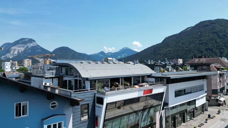 Moderne Architektur mit Glasfassade und Dachterrasse vor beeindruckender Bergkulisse und blauem Himmel.