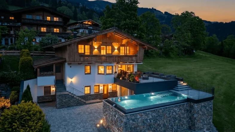 Luxuriöses Chalet in den Bergen bei Sonnenuntergang mit beleuchtetem Infinity-Pool und Terrasse.