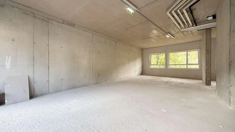 Großer, leerer Raum im Rohbauzustand mit Betonwänden und -decke sowie sichtbaren Installationen.