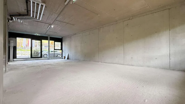 Großer, unfertiger Raum mit Betonwänden und -boden, bereit für den Ausbau. Große Fensterfront zur Straße hin.