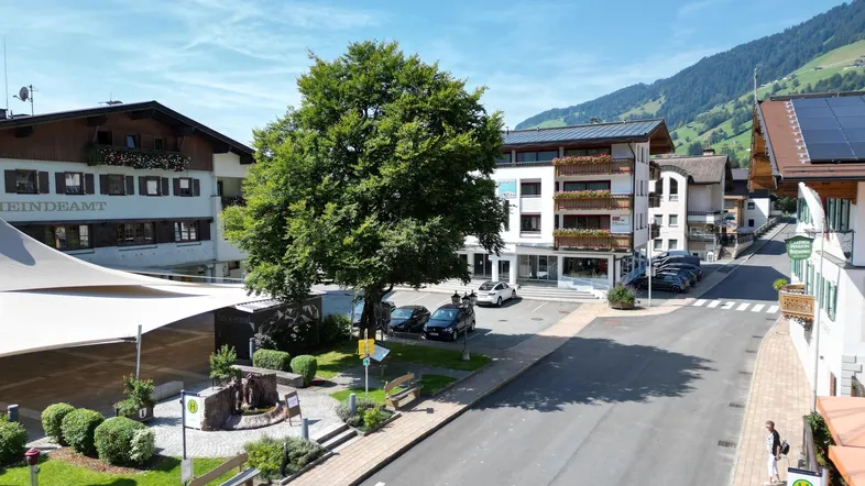 Luftaufnahme einer belebten Dorfstraße mit traditionellen Gebäuden, Bäumen und Bergen im Hintergrund unter blauem Himmel.
