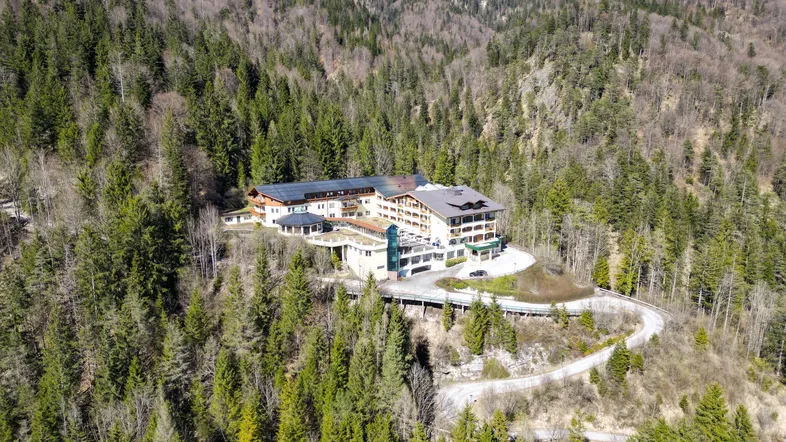 Luftaufnahme eines großen Gebäudekomplexes, eingebettet in eine bewaldete Berglandschaft unter blauem Himmel.