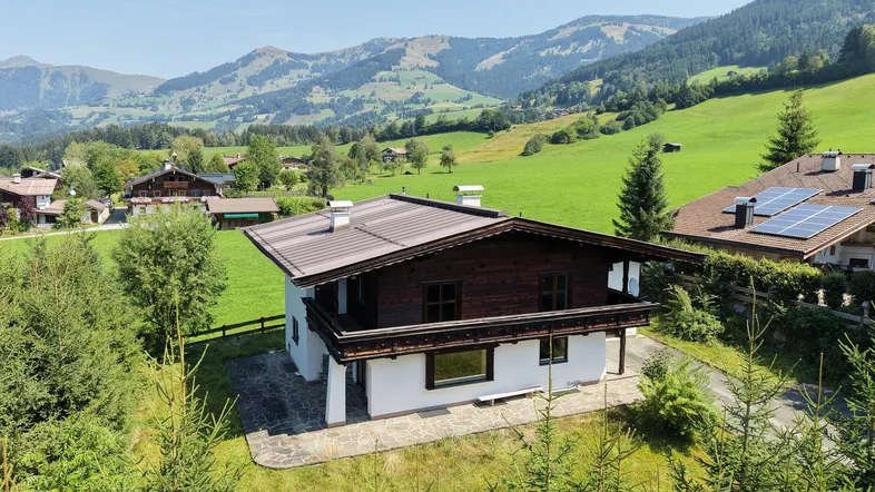 Haus im alpinen Stil mit Satteldach und Balkon, umgeben von grünen Wiesen und Bergen.