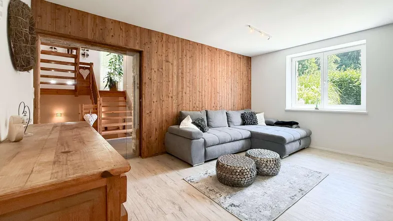 Helles Wohnzimmer mit Holzwand, modernem Sofa und Blick ins Grüne. Treppenzugang sichtbar.