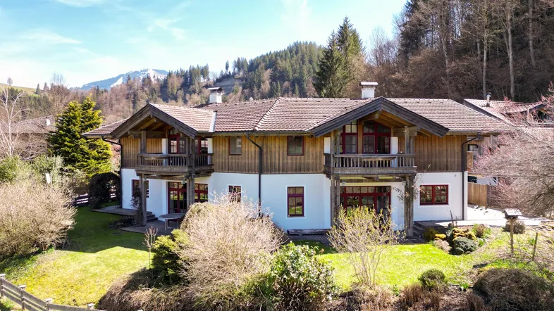 Traditionelles Landhaus mit Holzfassade und Satteldach, umgeben von grüner Natur und Bergen.