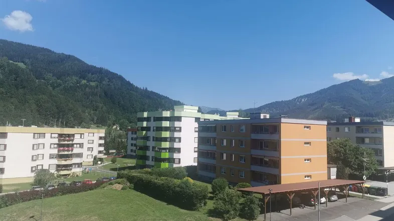 Blick auf mehrere Wohngebäude mit grünen Fassaden und die umliegende Berglandschaft unter blauem Himmel.