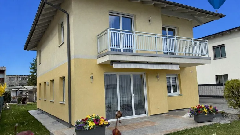 Gelbes Einfamilienhaus mit Balkon, Terrasse und gepflegtem Vorgarten unter blauem Himmel.