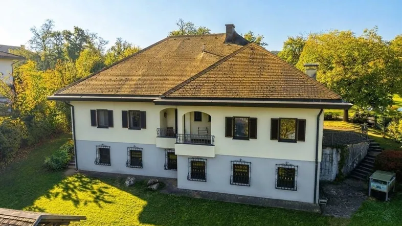 Traditionelles Haus mit heller Fassade, Satteldach und kleinem Balkon, umgeben von Grünflächen.