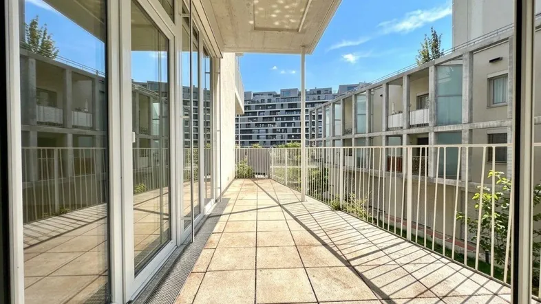 Langer Balkon mit Fliesenboden und weißem Geländer, bietet Blick auf moderne Wohngebäude.