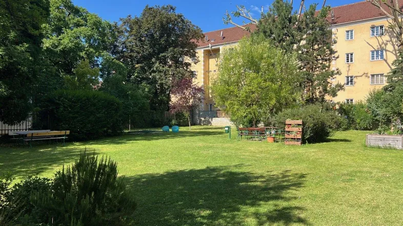 Grüner Gemeinschaftsgarten mit Rasenfläche, Bäumen und Sitzgelegenheiten vor einem Wohngebäude.