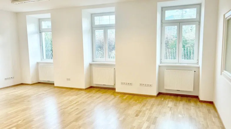 Heller, leerer Raum mit Holzparkettboden, drei großen Fenstern und Heizkörpern unter jedem Fenster.
