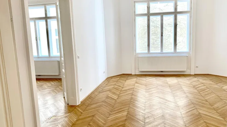 Heller Raum mit klassischem Fischgrätparkett, hohen Decken und großen Fenstern, der viel natürliches Licht bietet.