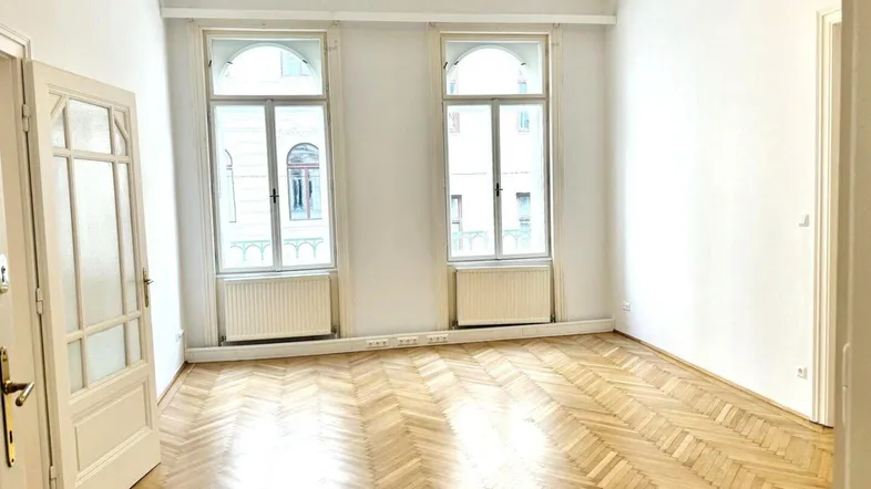 Helles Zimmer mit Fischgrätparkett, weißen Wänden und zwei großen Bogenfenstern.
