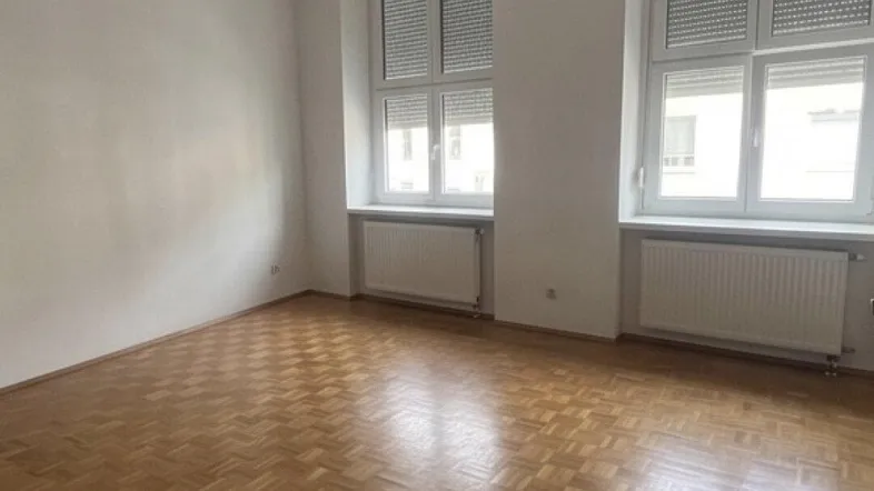 Helles, leeres Zimmer mit Parkettboden und zwei großen Fenstern, bereit zur individuellen Gestaltung.