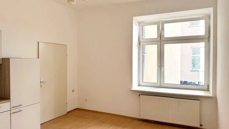 Helles Zimmer mit Holzboden, großem Fenster und Heizkörper, teilweise möbliert mit Küchenzeile.