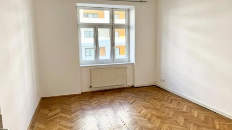 Helles Zimmer mit Fischgrätparkett, weißen Wänden und einem Fenster mit Blick nach draußen.