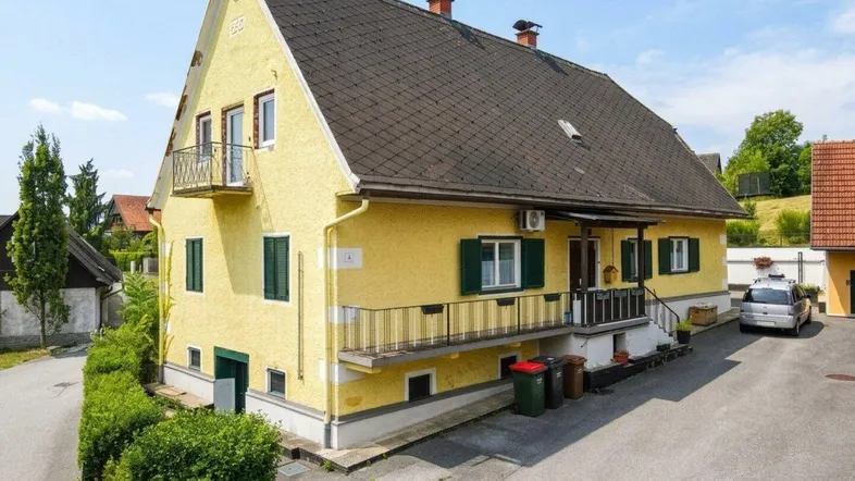 Gelbes Wohnhaus mit Satteldach, Balkonen und grünen Fensterläden in ländlicher Umgebung.
