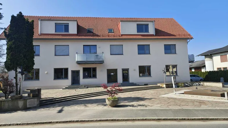 Mehrfamilienhaus mit heller Fassade, roten Dachziegeln und kleinem Balkon im Vordergrund.