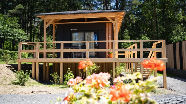 Modernes kleines Haus mit großer Holzterrasse und Blick ins Grüne, umgeben von Bäumen.
