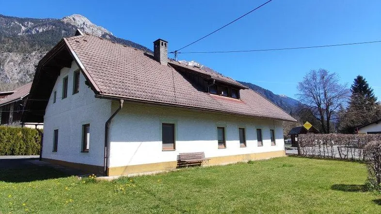 Ländliches Haus mit Satteldach und großem Garten in bergiger Umgebung unter blauem Himmel.