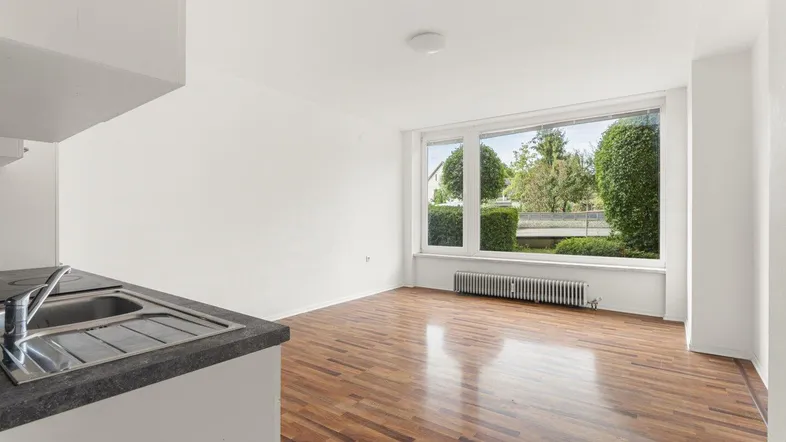 Helles Wohnzimmer mit Holzboden, großem Fenster und Blick ins Grüne, inklusive kleiner Küchenzeile.