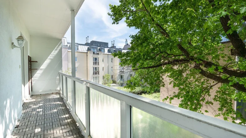 Geräumiger Balkon mit Holzbodenfliesen und Blick ins Grüne sowie auf umliegende Gebäude.