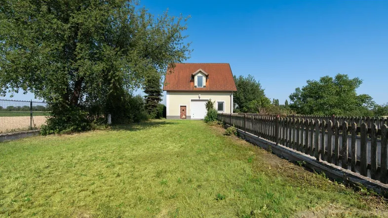 Kleines Haus mit rotem Dach und hellem Anstrich, umgeben von einem großen Garten und Bäumen.