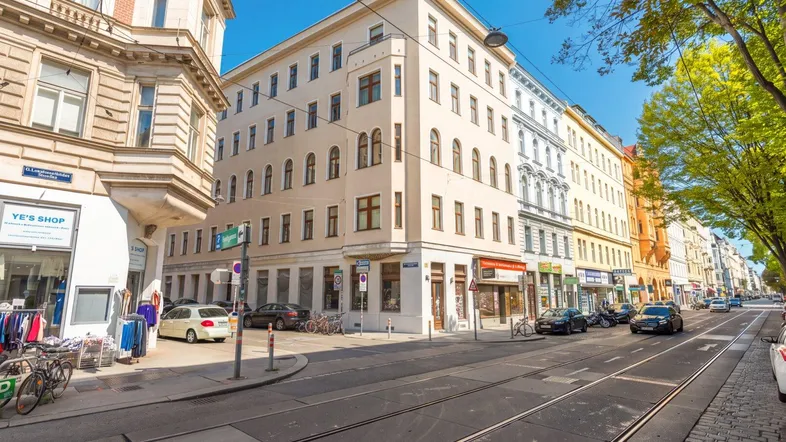 Historisches Eckgebäude in einer belebten Stadtstraße mit Geschäften und Straßenbahnschienen unter blauem Himmel.