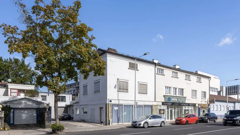 Weißes Mehrfamilienhaus mit Geschäftsflächen im Erdgeschoss und Parkplätzen an der Straße.
