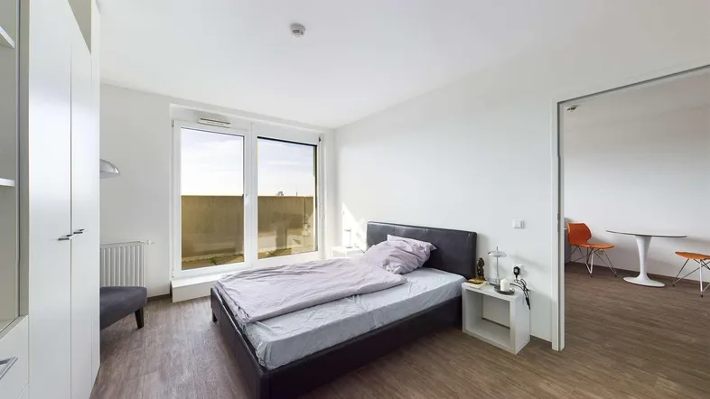 Helles Schlafzimmer mit Holzboden, großem Fenster und Zugang zu einem weiteren Raum.