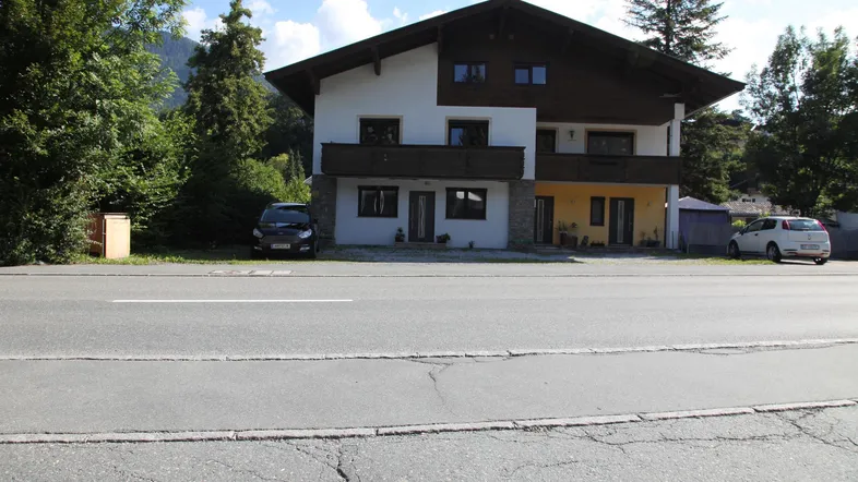 Traditionelles Haus mit Satteldach und Balkon, umgeben von Bäumen und Bergen.