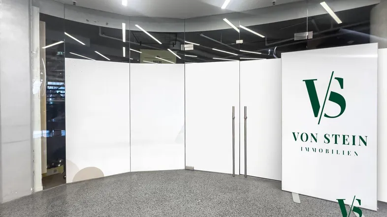 Eingangsbereich mit geschwungenen Glaswänden, modernem Terrazzoboden und Immobilienwerbung.