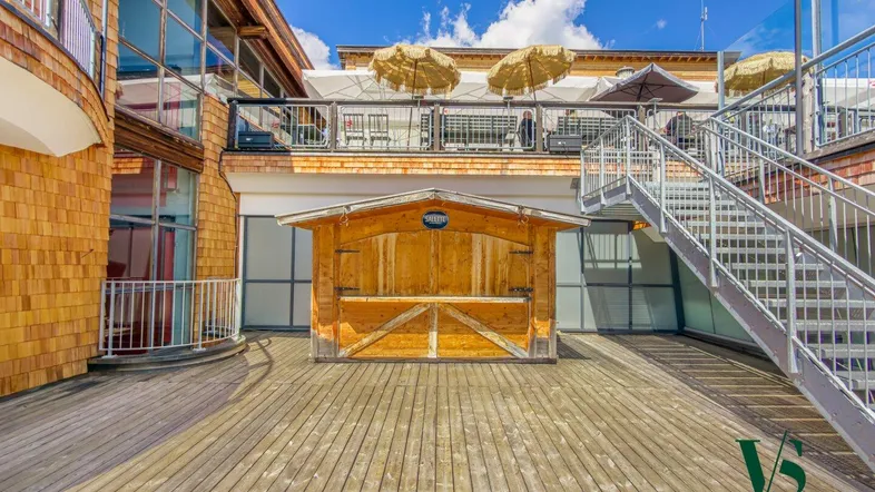 Weitläufige Holzterrasse vor einem modernen Gebäude mit großen Glasflächen und einem kleinen Holzstand.